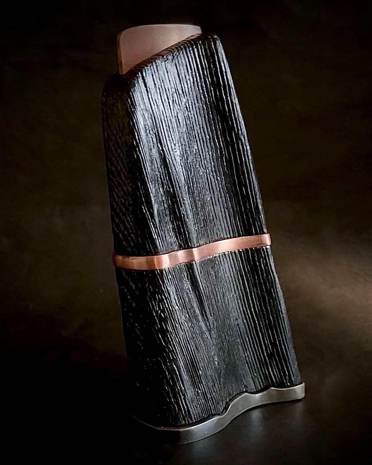 A free-form black sculpture with a copper band, displayed against a dark background.