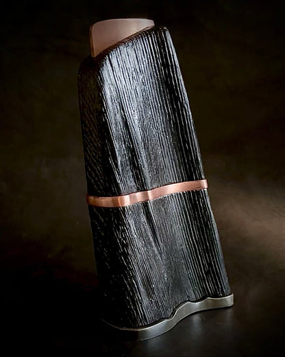 A free-form black sculpture with a copper band, displayed against a dark background.