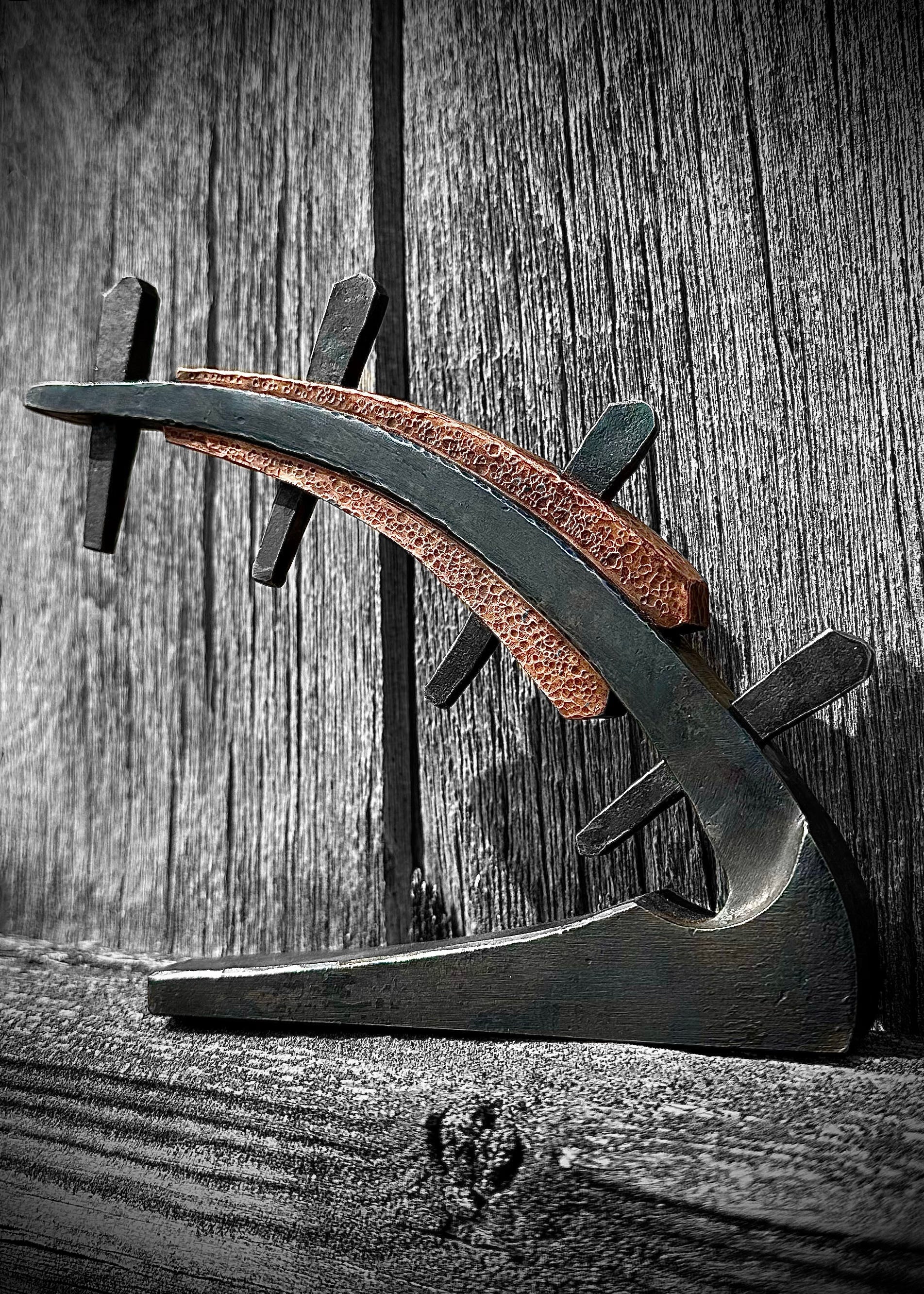 A sculpture with a curved form, featuring a hammered copper element and forged steel markers, placed against a wooden background.