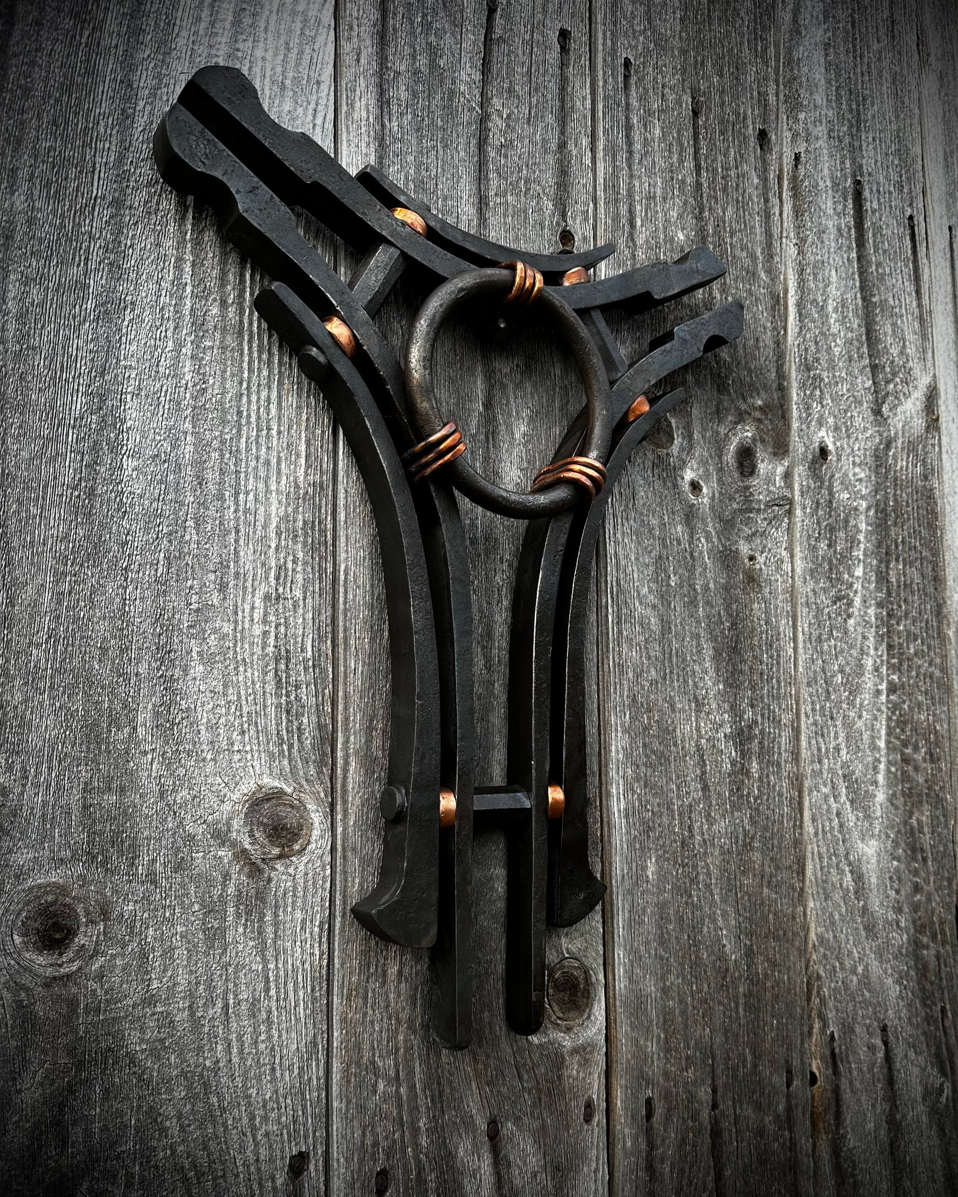 A hand-forged sculpture of steel and copper with a central ring and three interconnected arms, displayed against a rustic wooden background.