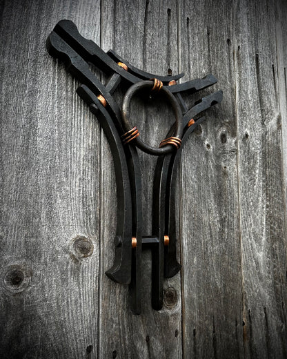 A hand-forged sculpture of steel and copper with a central ring and three interconnected arms, displayed against a rustic wooden background.