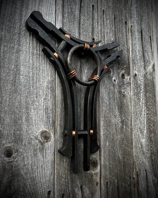 A hand-forged sculpture of steel and copper with a central ring and three interconnected arms, displayed against a rustic wooden background.