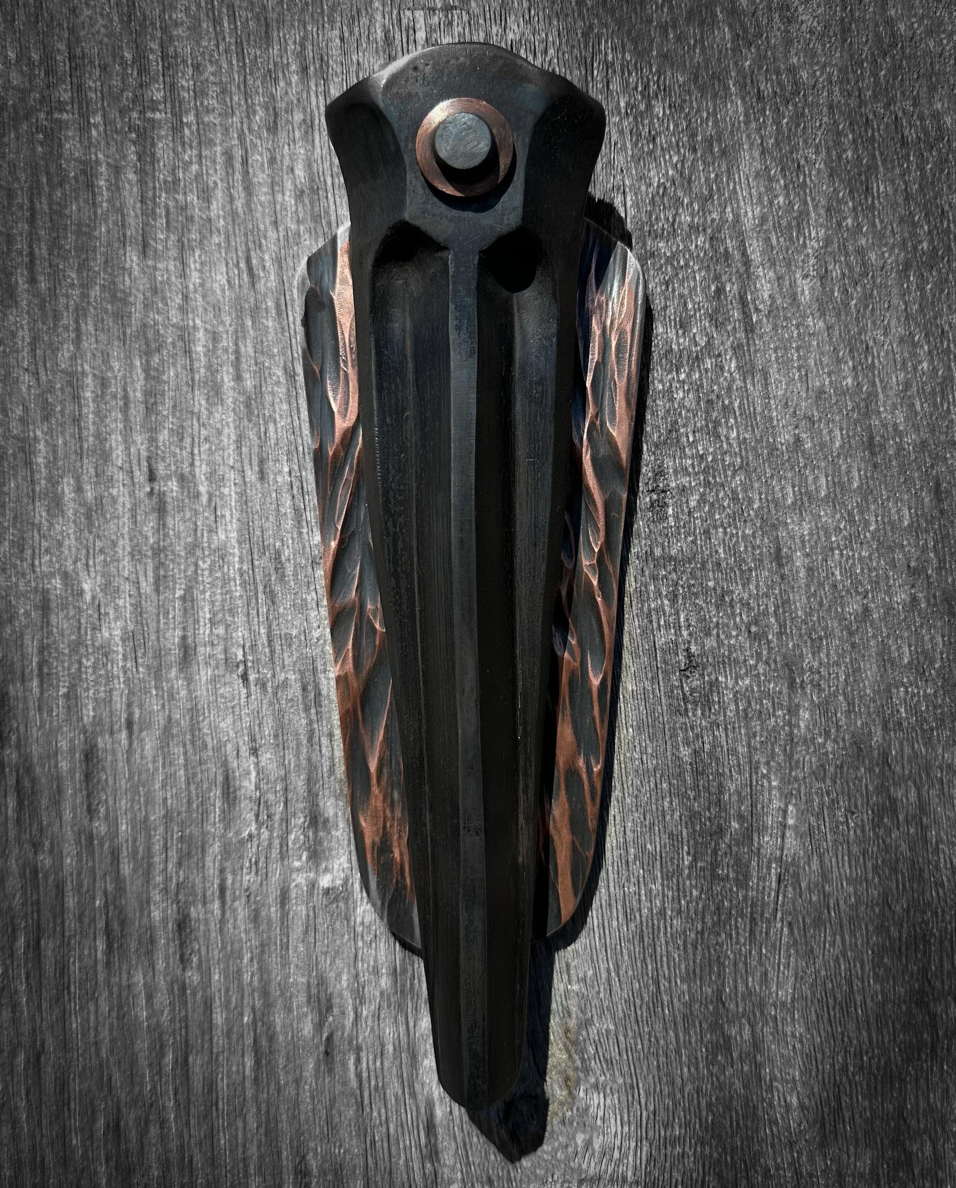 A vertical sculpture made from dark steel and copper, with the copper elements having a textured, patinated finish. The sculpture appears abstract and minimalist, displayed against a grey background.