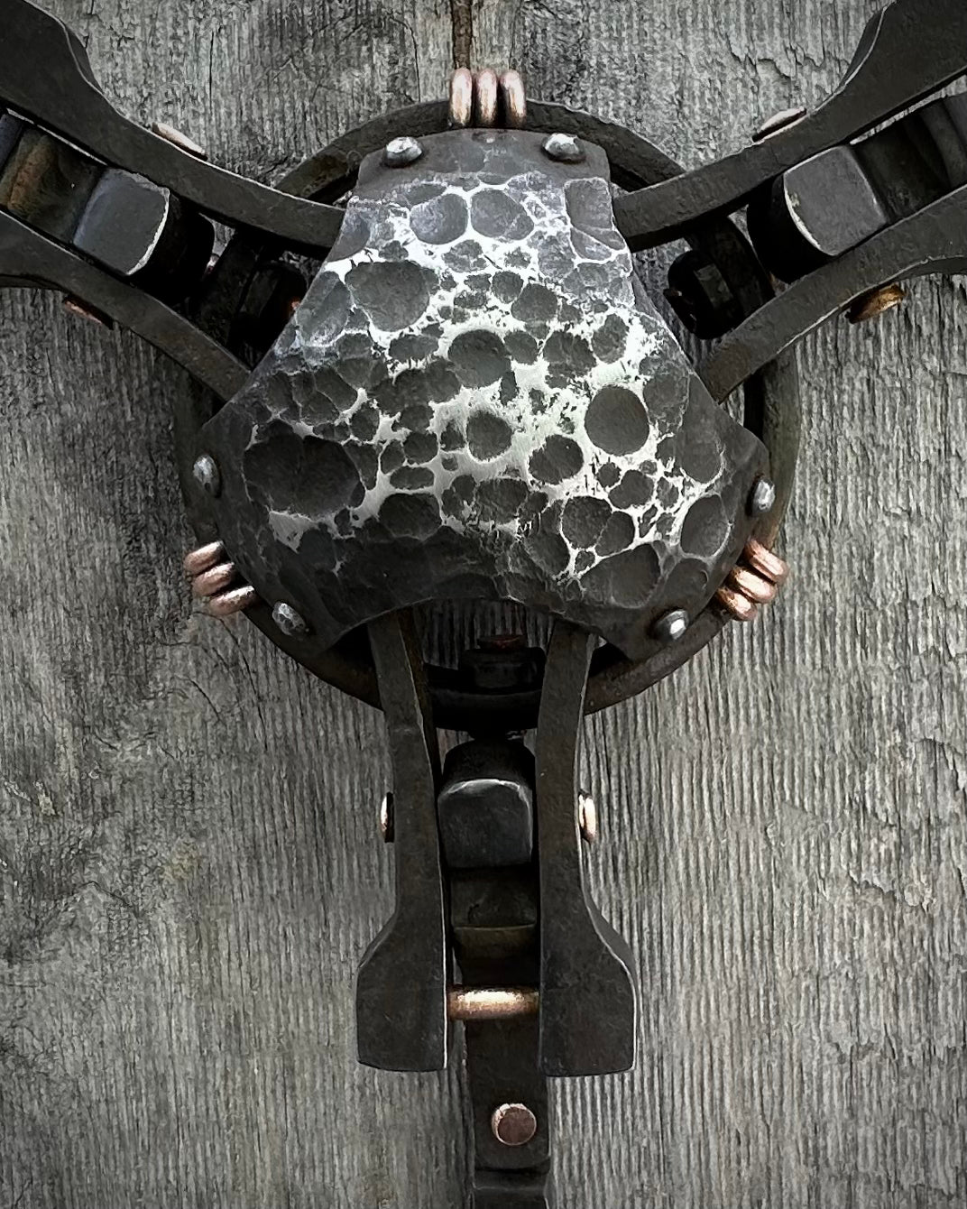 A close-up of a textured sculpture made of steel and copper with a patina finish, featuring a mix of smooth and rough surfaces, mounted on a wall.